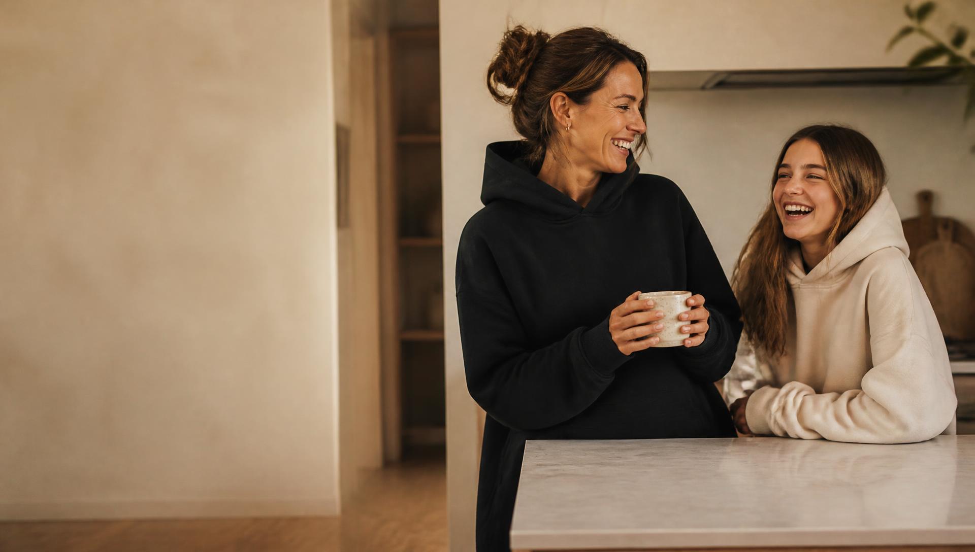 A mother and her teenage daughter laughing in a sunlit kitchen, both in oversized MARVAB hoodies